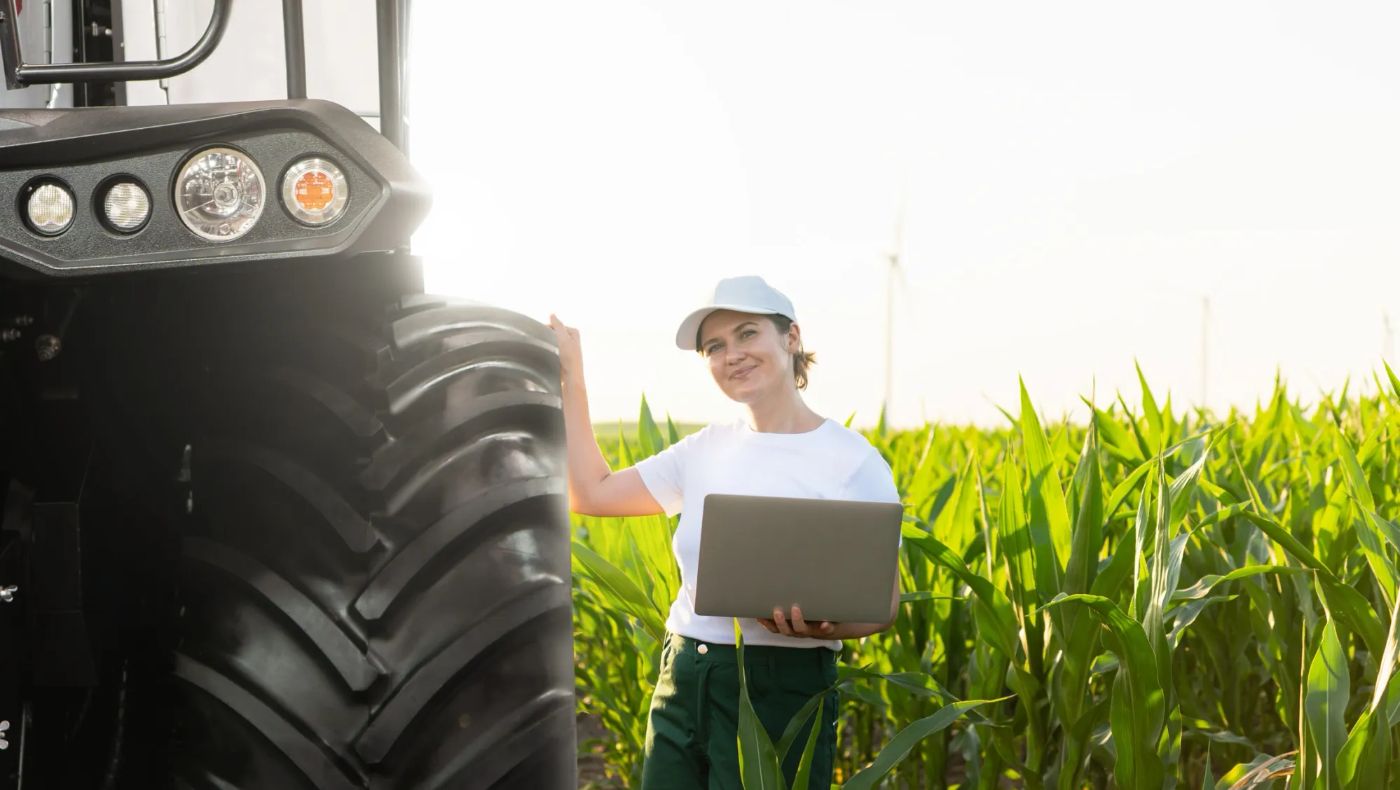 Produtora rural com notebook ao lado de trator em plantação